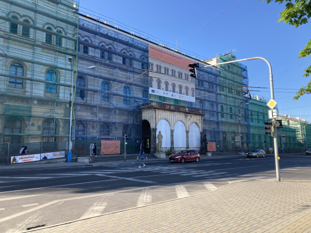 Großes Gebäude mit Gerüst während der Renovierung, umgeben von Fahrzeugen, Fußgängern, Ampeln, Schildern, Straßeninfrastruktur, Bäumen und einem klaren blauen Himmel.