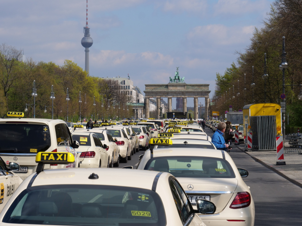 Eine belebte Straße in Berlin mit zahlreichen geparkten Taxis, Fußgängern auf dem Gehweg, gesäumt von Laternenmasten und Bäumen, Gebäude im Hintergrund, ein fernes Tor mit Statuen und Turm und ein bewölkter Himmel.