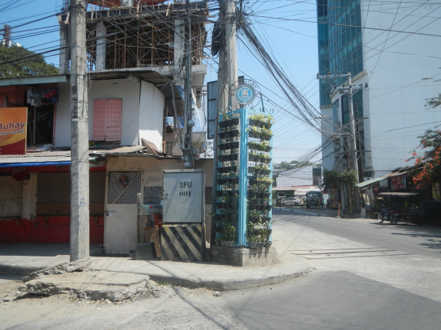 Stadtstraße mit Gebäuden, Strommasten, Kabeln, Namensschildern, Fahrzeugen, Bäumen und einem Gebäude mit Drähten im Vordergrund unter einem Himmel.