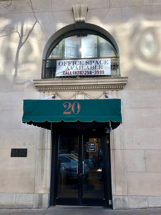 Gebäude mit einer Glastür, grüner Markise, einem Schild mit der Aufschrift "Büroräume zu vermieten", einem Geländer und einem Baum auf der linken Seite.