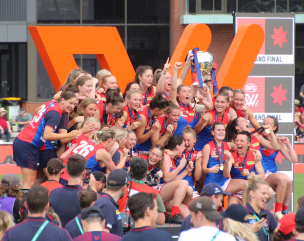 Gruppe von Frauen-AFL-Spielerinnen, die mit einem Pokal vor einem Publikum feiern, tragen Sportuniformen, einige halten den Pokal, mit einem Gebäude und einer Tafel im Hintergrund.