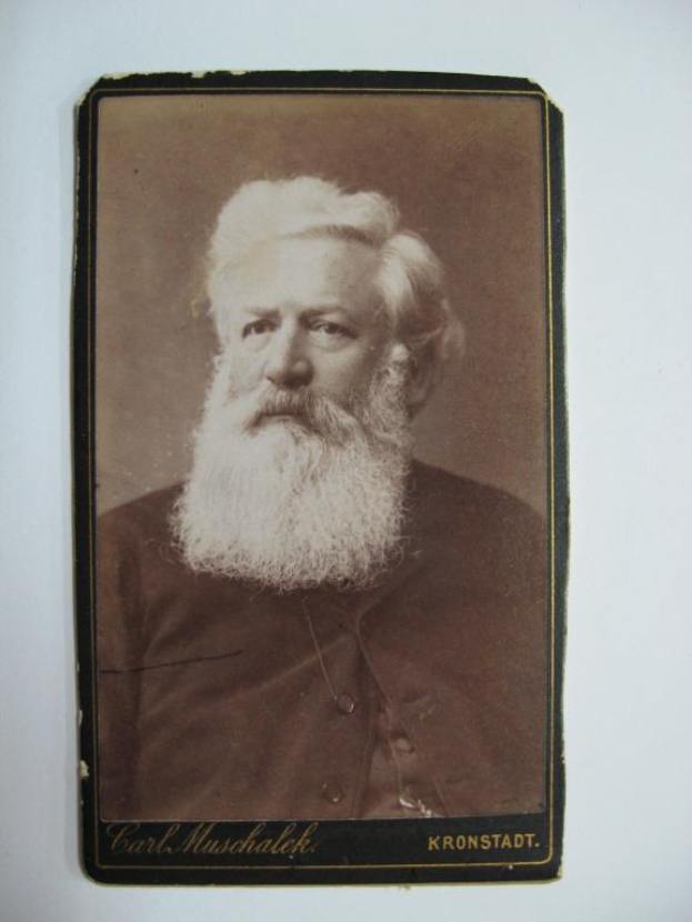 Schwarz-weiß-Foto von Carl Muschalk, einem deutschen Komponisten mit langem weißem Bart, an einer weißen Oberfläche befestigt mit Text unten.