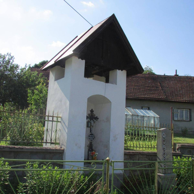 Kleines weißes Gebäude mit einem Kreuz darauf, umgeben von einem Metallzaun, in einem Gebiet mit Pflanzen, Gras, einem Weg, Häusern, Pfählen, Drähten und einem bewölkten Himmel.