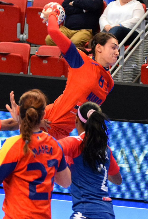 Eine Gruppe von Frauen beim Handballspielen in einem Stadion, mit einer Spielerin, die den Ball hält und Zuschauern im Hintergrund.