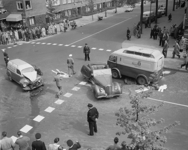 Schwarz-weiß-Szene eines Autounfalls an einer Straßenecke mit mehreren Fahrzeugen, darunter ein Lieferwagen, und einer Gruppe von Schaulustigen; Gebäude, Bäume, Strommasten und Texttafeln im Hintergrund.