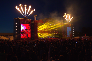 Eine große Menschenmenge auf einem Musikfestival mit einer Bühne, auf der Künstler performen, helle Lichter und Banner, Zelte links daneben und Feuerwerk am dunklen Himmel.