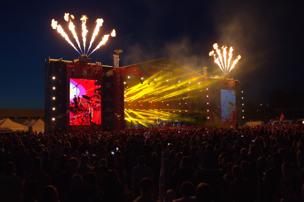 Eine große Menschenmenge auf einem Musikfestival mit einer Bühne, auf der Künstler performen, helle Lichter und Banner, Zelte links daneben und Feuerwerk am dunklen Himmel.