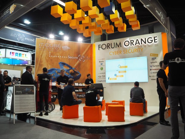 Eine Gruppe von Menschen an einem Messestand für IT-Cube Systems Forum Orange Cyber Security, mit sitzenden und stehenden Teilnehmern, einem Anzeigebildschirm, Regalen mit Gegenständen, Schildern und Deckenleuchten.
