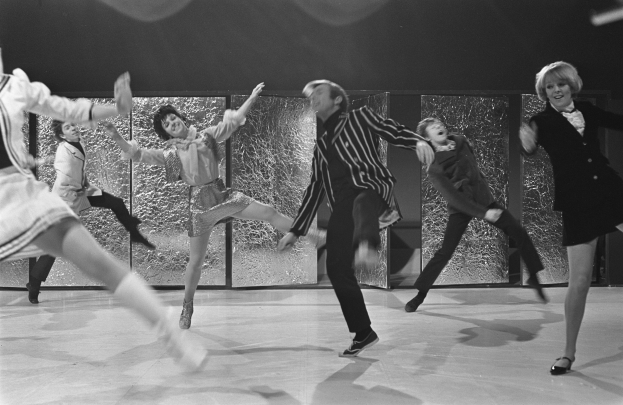 Eine Gruppe von Menschen, die auf einer Bühne tanzen, vor einer Wand, in Schwarz-Weiß, alle tragen Schuhe.