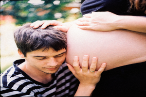 Ein Mann und eine Frau stehen nebeneinander, der Mann beruhrt zart den Bauch der Frau, der Hintergrund ist unscharf.