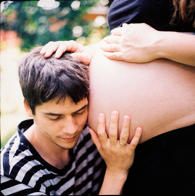 Ein Mann und eine Frau stehen nebeneinander, der Mann beruhrt zart den Bauch der Frau, der Hintergrund ist unscharf.