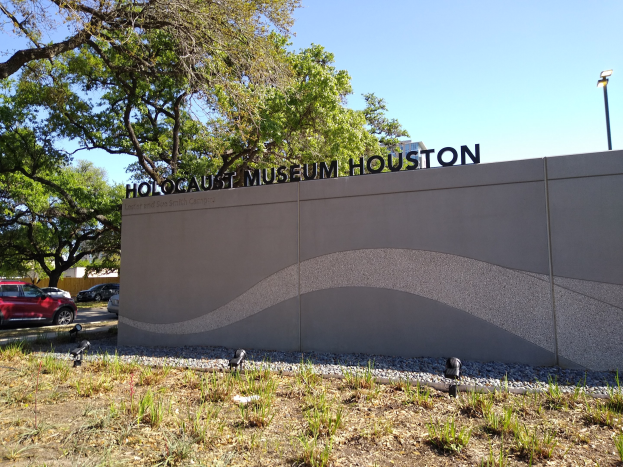Außenansicht des Houston Holocaust Museums mit einer Gedenkwand, Steinen, Vegetation, Fahrzeugen, einem Straßenschild und einem bewölkten Himmel.