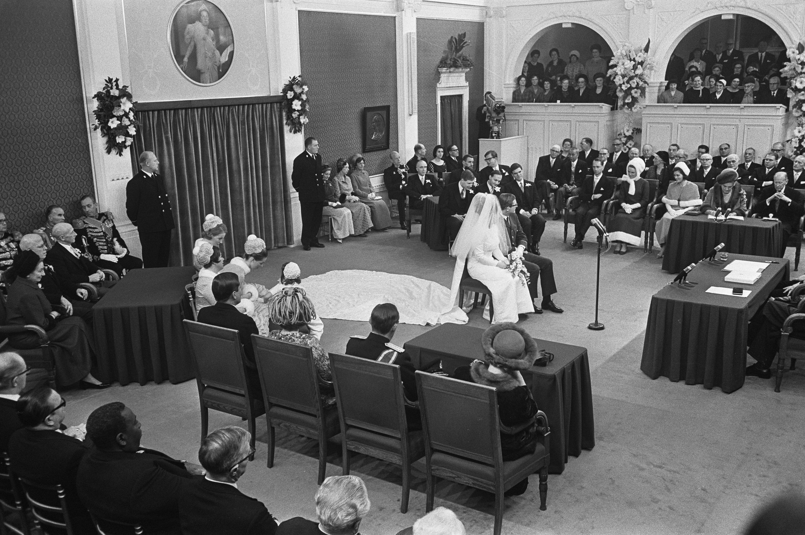 Schwarz-weiß-Foto einer Hochzeitszeremonie in einem großen Raum mit sitzenden und stehenden Gästen, Tischen mit Papieren und Mikrofonen, einer Wand mit Rahmen, Blumenbouquets, einem Vorhang und einem Podium mit Mikrofonständer.