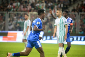 Ein Fußballspieler in einem blauen und weißen Trikot feiert mit erhobenen Armen, umgeben von einer Gruppe in weißen und grünen Trikots, mit einem unscharfen Hintergrund, der ein weißes und blaues Brett und ein Geländer zeigt.