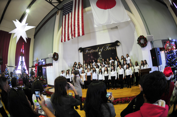 Eine Gruppe von Menschen auf einer Bühne mit Weihnachtsdekoration, einem Rednerpult, Lautsprechern, einer Texttafel und einer Menge darunter mit Kameras und Handys.