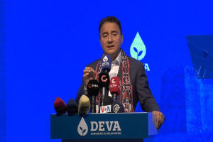 Ein Mann in einem Mantel und Schal steht an einem Podium mit Mikrofonen, ein Schild mit der Aufschrift "deva-Geschäftsführer sagt, es ist keine gute Idee für die Zukunft", ein Objekt und ein Bildschirm im Hintergrund.