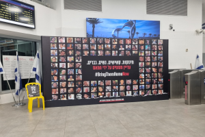 Eine große Wand zeigt zahlreiche Bilder, einen gelben Stuhl, israelische Regierungsflaggen auf Ständern, eine Tafel mit der Aufschrift "Bring Them Home Now", einen montierten Bildschirm, Deckenleuchten und andere Gegenstände.