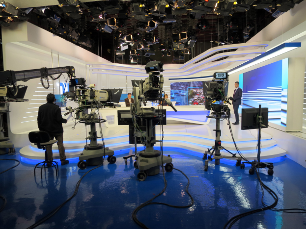 Ein TV-Studio mit mehreren bodengestützten Kameras, einer sitzenden Person im Vordergrund, stehenden Crew im Hintergrund und deckengestützten Lichtern, die eine Live-Sendung andeuten.