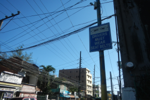 Stadtstraße mit fahrenden Autos, Strommasten mit Überlandleitungen, Gebäude mit Fenstern, Bäume, Schilder und ein "Fahrer müssen Sitzgurte tragen"-Schild an einem Strommasten vor einem sichtbaren Himmel.