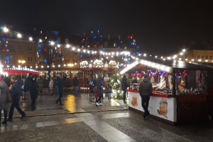 Ein belebter Weihnachtsmarkt bei Nacht in einer Stadt mit Menschen, die herumlaufen, Ständen mit Lichtern und Dekorationen und Gebäuden mit Fenstern und Bäumen im Hintergrund.