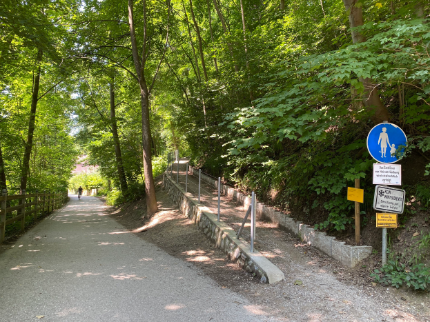 Eine Person fährt ein Fahrrad auf einem asphaltierten Weg durch den Wald, mit Bäumen und einem Holzzaun auf der linken Seite und Schildern mit Text und Bildern auf der rechten Seite.