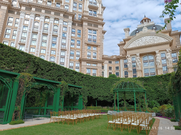 Hochzeitszeremonie im Freien mit Stühlen, Pavillon und Blumenschmuck vor einem Glasgebäude unter einem blauen Himmel.