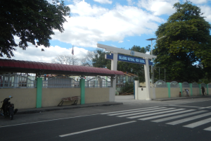 Einfahrt zu einer Schule mit einem vor der Tür geparkten Motorrad, einer Straße mit weißen Linien und Zebrastreifen, einer Mauer mit einem Schuppen, einem Schild, Laternen, Bäumen, einer Fahne auf einem Mast und einem bewölkten Himmel.