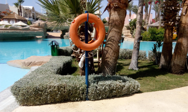 Schwimmbad mit einem Rettungsring in der Mitte, umgeben von grünem Gras, Pflanzen, Bäumen, Gebäuden, Sonnenschirmen und einem klaren blauen Himmel.