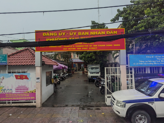 Ein Polizeiwagen steht vor einem Gebäude mit der Aufschrift "Dang Uy Uy Ban Nhan Dan". Im Hintergrund sind verschiedene Gebäude, Fahrzeuge und ein Turm zu sehen.