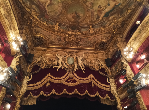 Das Innere eines Theaters mit einem roten Vorhang, Kronleuchtern, Skulpturen, einer Deckenmalerei und einer Bühne mit einigen Menschen davor.