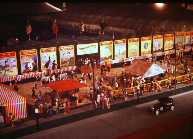 Ein altes Faschingsbild mit Menschen, einem Auto, Masten mit Fahnen, einem Zaun, einem Zelt, Brettern mit Bildern und Text sowie einer Laterne.