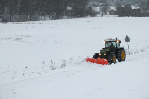 Eine Person fährt einen Traktor mit einer Schneepflug durch ein schneebedecktes Feld, mit Bäumen, Häusern und einem klaren blauen Himmel im Hintergrund.