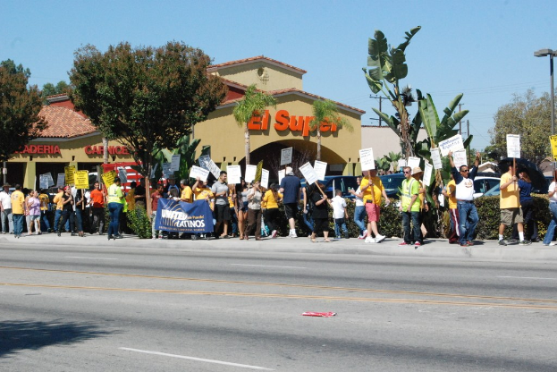 Eine Gruppe von Menschen mit Schildern, die auf einer Straße mit kleinen Büschen, einer Fahne, einem Gebäude mit Namensschildern und Autos im Hintergrund gehen.