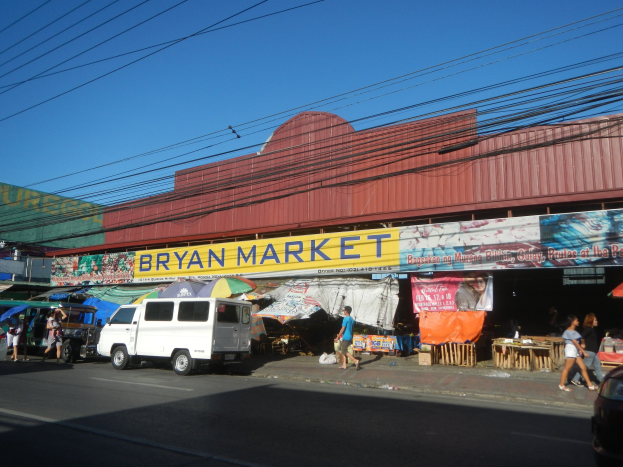 Eine belebte Marktplatzszene mit Fahrzeugen, Fußgängern, Marktständen, Bannern, Schirmen, hölzernen Gegenständen und einem "Bryan Market"-Gebäude im Hintergrund unter einem Himmel mit Oberleitungen.