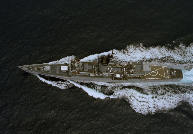 Eine lenkwaffenbestückte Fregatte segelt auf dem Ozean, wobei sich ihr Rumpf in der Sonne spiegelt und Wellen an ihren Seiten lecken, mit einem prominenten Geschützturm und blähenden Segeln.