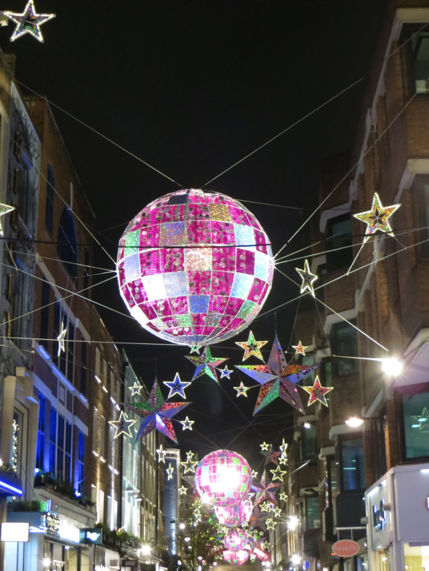 Eine festliche Stadtstraße bei Nacht mit zahlreichen Lichtern und Dekorationen, darunter eine große Diskokugel an einem Gebäude.
