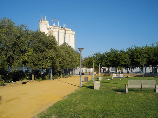 Ein Park mit Bänken und Bäumen vor einem Gebäude, umgeben von Straßenmasten, Straßenlaternen, Grills, Pflanzen und einem klaren blauen Himmel.