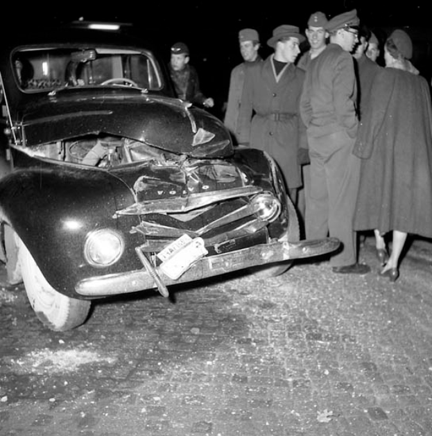 Eine schwarz-weisse Szene eines beschädigten Autos am Strassenrand, mit einer Gruppe von Menschen drumherum, die das Auto besorgt anschauen.