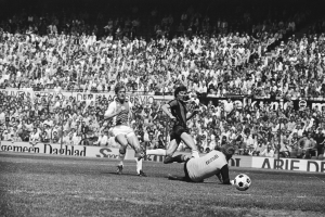 Schwarzes und weißes Foto von Fußballspielern im Einsatz auf einem Feld mit Zuschauern im Hintergrund und einer "Bundesliga 1978-79 Ajax 1-1 Hannover 96"-Fahne.