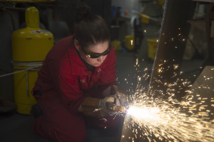 Eine Frau in einem roten Kleid und Schutzbrille schweißen Metall in einer Fabrik, mit Funkenflug und industrieller Ausrüstung im Hintergrund.