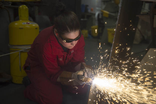 Eine Frau in einem roten Kleid und Schutzbrille schweißen Metall in einer Fabrik, mit Funkenflug und industrieller Ausrüstung im Hintergrund.