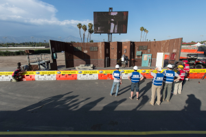 Eine Gruppe von Menschen in Helmen steht vor einem großen Bildschirm auf dem Las Vegas Motor Speedway, umgeben von Barrieren, Fahrzeugen, Hütten, Bäumen, Lichtmasten und Bergen mit bewölktem Himmel im Hintergrund.