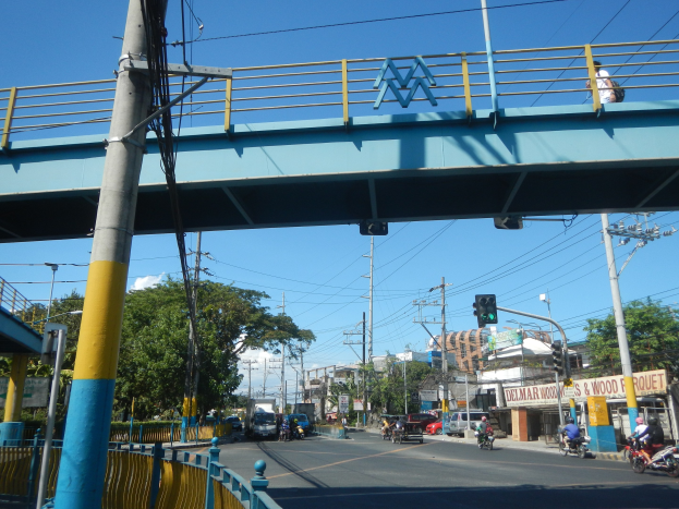 Fußgängerbrücke über eine vielbefahrene Straße mit Fahrzeugen, Strommasten, Verkehrsampeln, Geländer, Bäumen, Gebäuden und Himmel im Hintergrund.