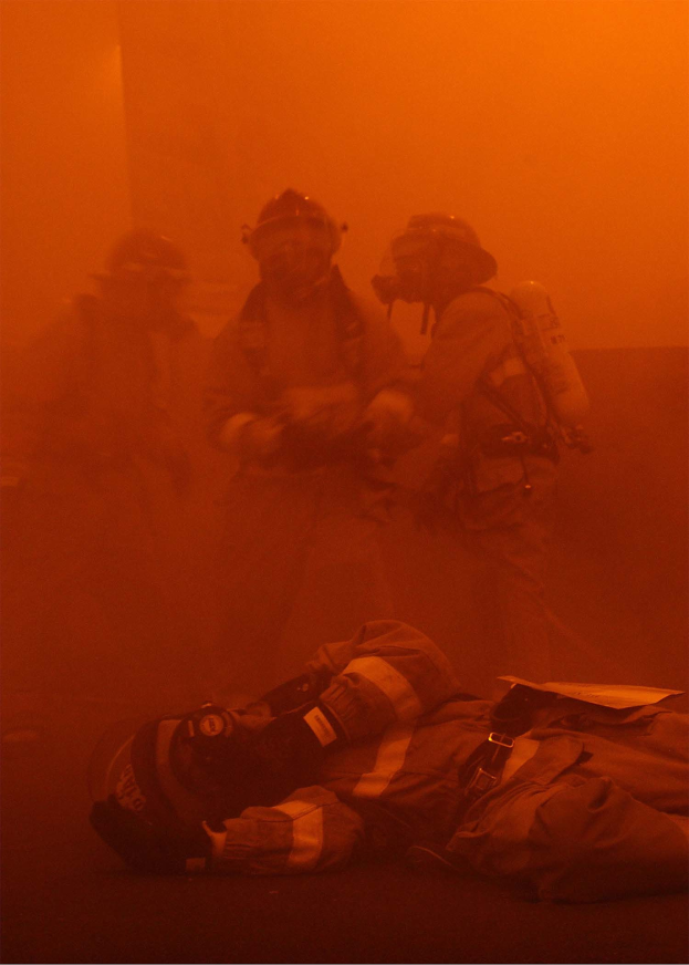 Schwarz-weißes Bild eines Feuerwehrmanns, der vor intensiven Flammen auf dem Boden liegt, umgeben von einer Gruppe von Menschen mit Helmen und Sauerstofftanks.