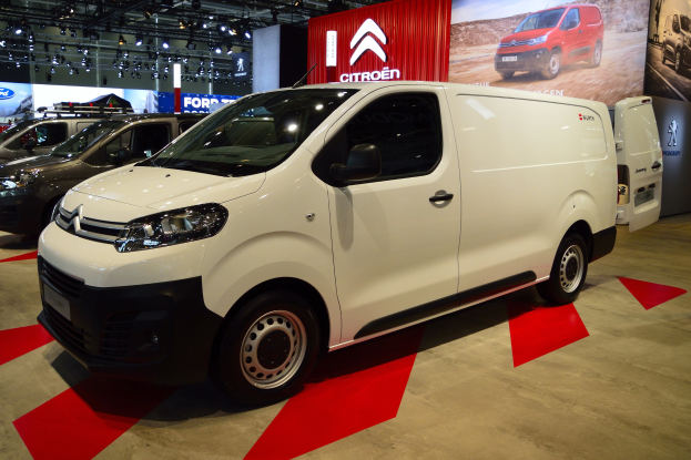 Weißer Citroen Berlingo Van zwischen schwarzen Schlafzimmermöbeln auf einer Autauschau, mit beleuchteten Wandtafeln für eine moderne, stylische Atmosphäre.